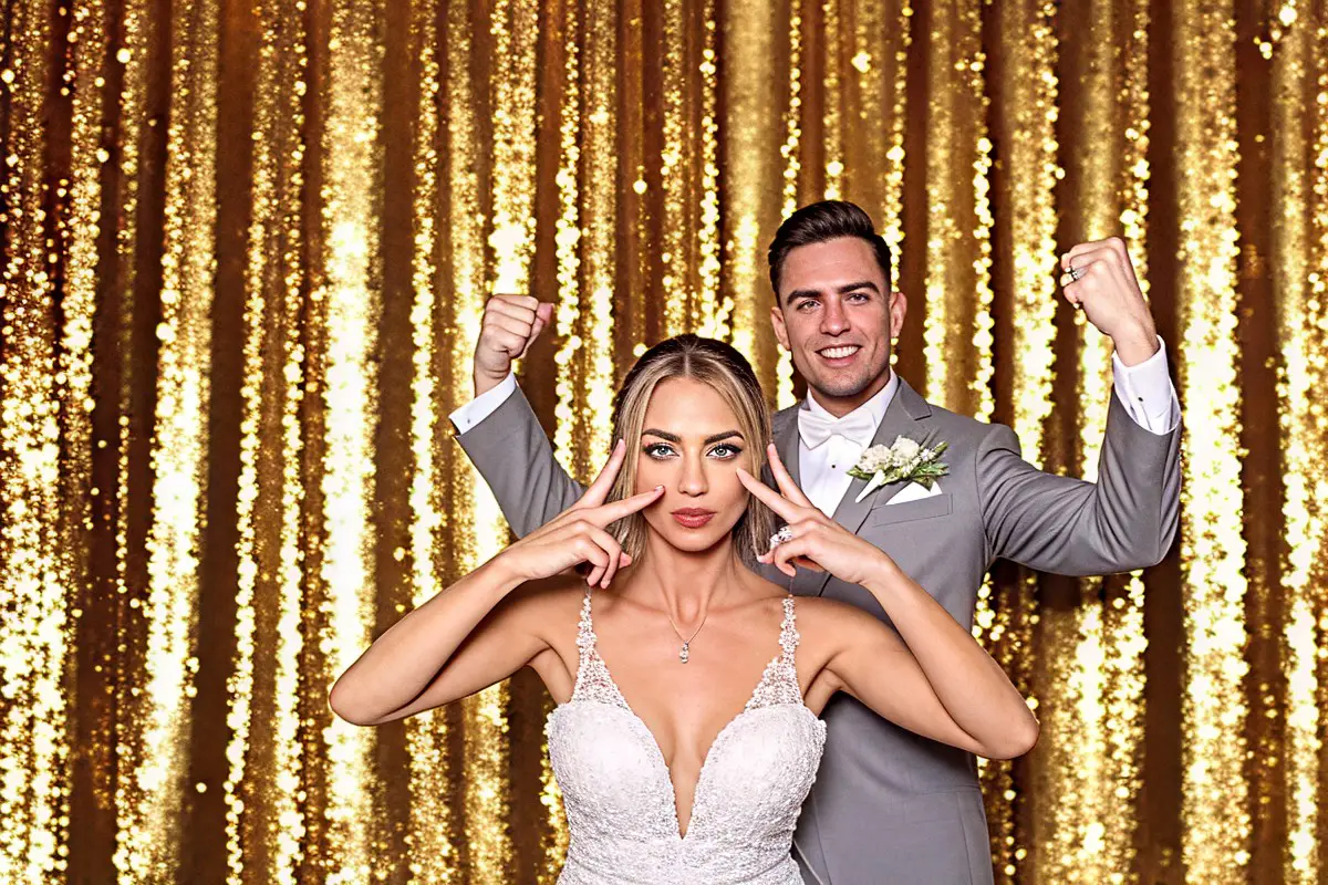 Hochzeitspaar posiert vor goldenem Pailettenvorhang, Braut zeigt Peace-Zeichen, Bräutigam zeigt Bizeps, Fotospiegel auf Hochzeit in Pforzheim